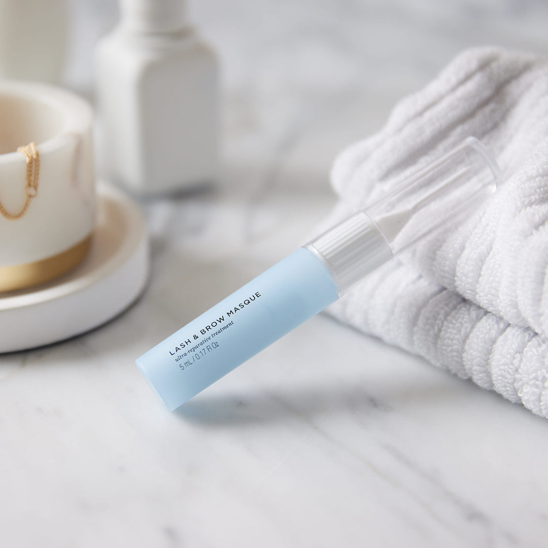 Image of a tube of Lash & Brow Masque leaning against white hand towels on a countertop